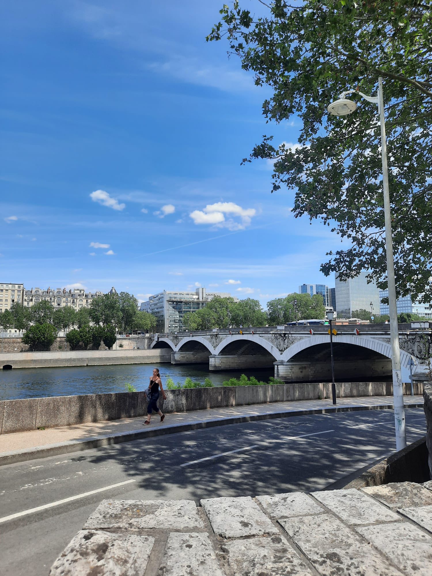 709664620_1290075479946942_5974212703341303333_n Woman walking along a riverside promenade with a stone wall, arched bridge over water, and city buildings in the distance under a blue sky.
