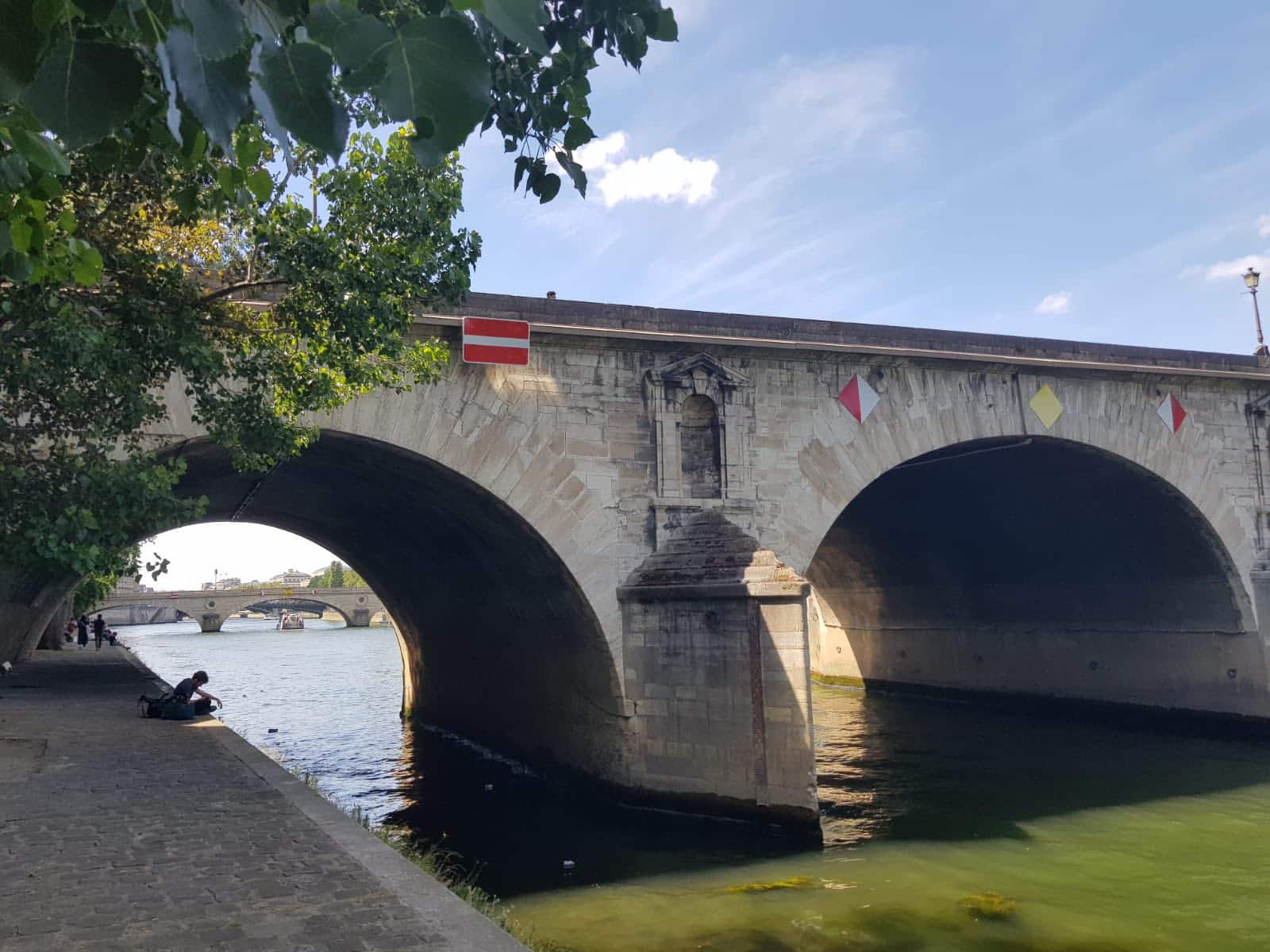 709664620_1290075489946941_9118853133187655067_n Stone arched bridge over a calm river with trees along the bank and a red no-entry sign on the side.