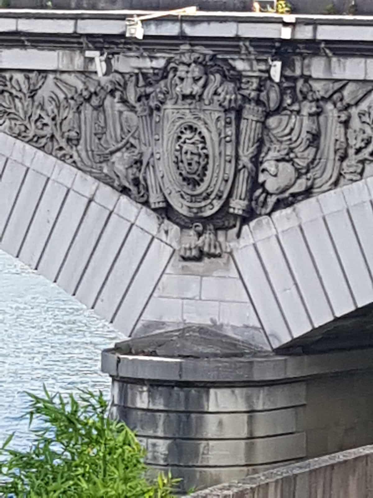 709881413_1290074769947013_1344867435528683140_n Ornate stone relief with a central shield motif and decorative figures on a bridge arch over water, in grayscale.