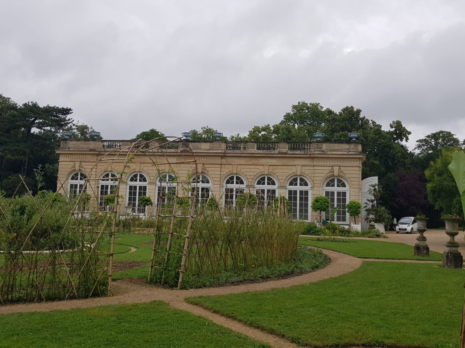 710059078_1290691733218650_8927831429973618987_n Beige neoclassical villa with five arched windows, set in a formal garden with gravel paths and climbing trellises on a cloudy day.