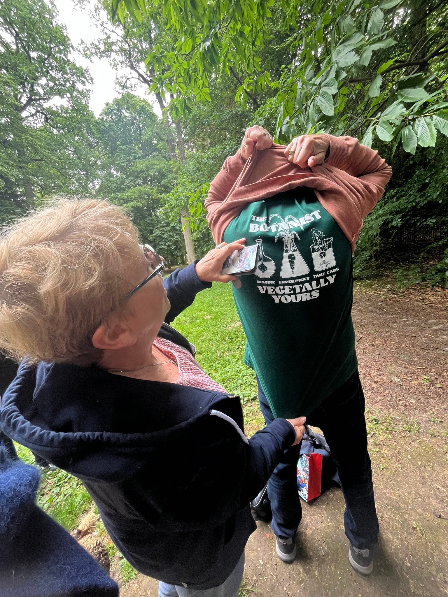 710252293_1291534759801014_3231445924159939460_n Two people outdoors in a park; one lifts a jacket to reveal a green T‑shirt with science-themed graphics reading 'The Botanist' and beakers.
