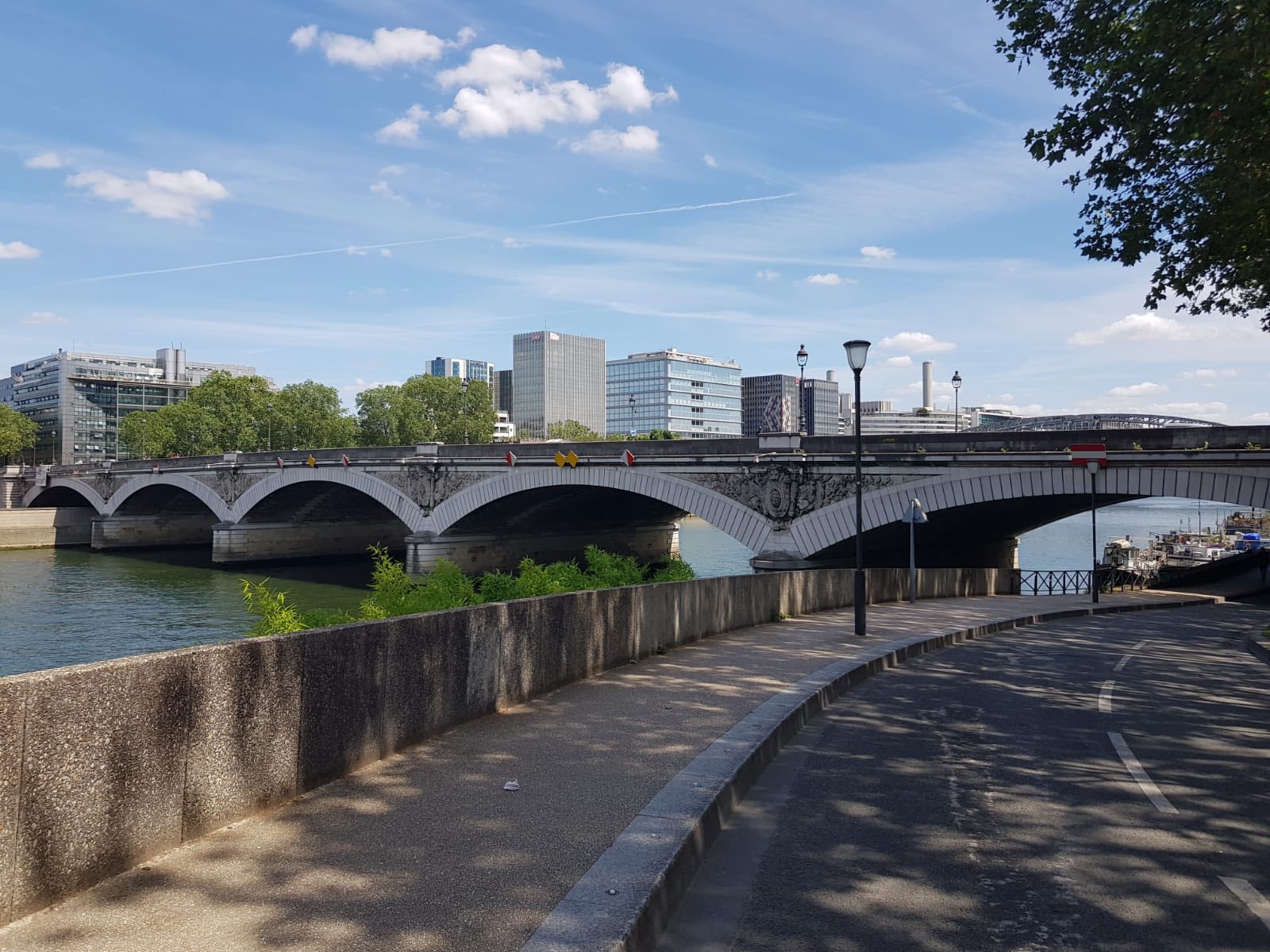 710279773_1290075039946986_841559050947754293_n Stone arched bridge crossing a calm river with modern office buildings and a blue sky behind it, a riverside walkway in the foreground.
