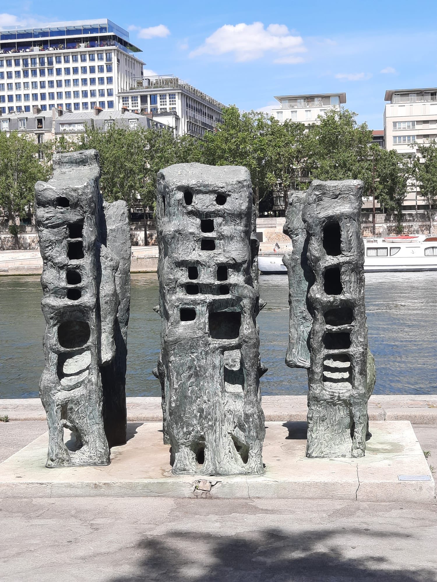 710306916_1290076256613531_3982176720018350318_n Group of tall stone sculpture columns with rectangular cutouts along a riverside promenade.