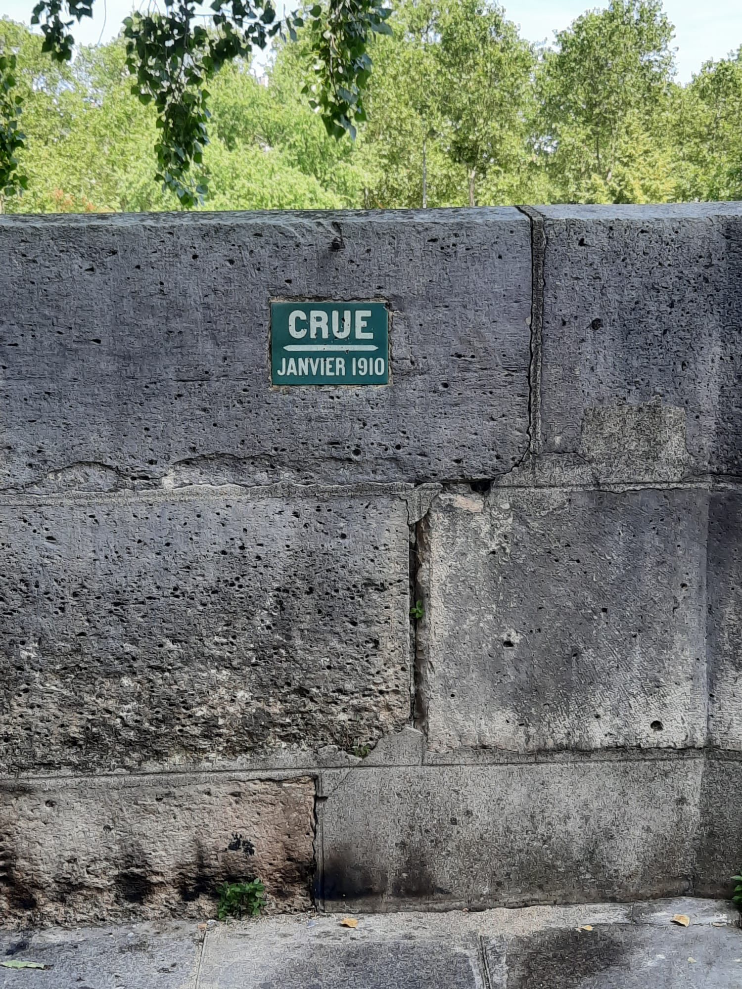 710461096_1290074656613691_584095966633961624_n Green plaque on a weathered concrete wall reading 'CRUE JANVIER 1910' with a crack and some greenery at the bottom; trees visible in the background.
