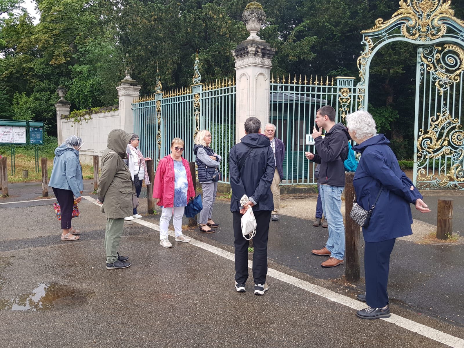 710596585_1290692376551919_3089681084007192739_n Tour group standing by an ornate iron gate with gold accents, listening to a guide on a cloudy day.