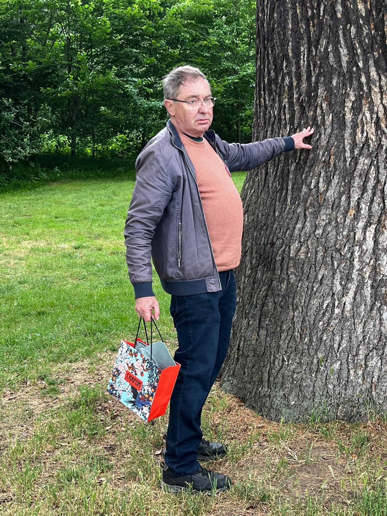 710700625_1291533393134484_8984529956643652186_n Older man in a peach sweater and gray jacket stands beside a large tree, touching the trunk in a grassy park, carrying gift bags.