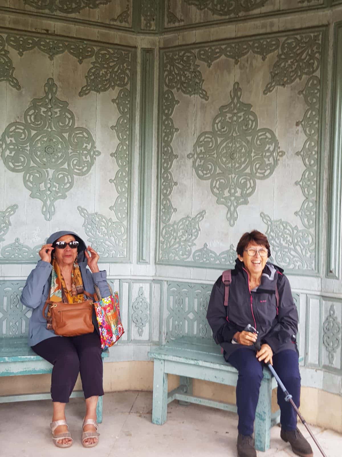 712045309_1290691313218692_5867979630105952317_n Two smiling women sit on a turquoise carved bench inside a decorative pavilion, one adjusting sunglasses and carrying a colorful bag.