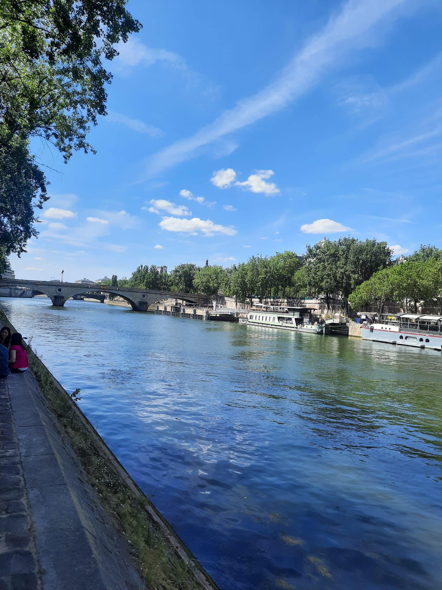 712445139_1290076219946868_2018538599647681989_n River scene with a stone arch bridge crossing calm water; trees line the riverbank and a walkway on the left under a blue sky with scattered clouds.