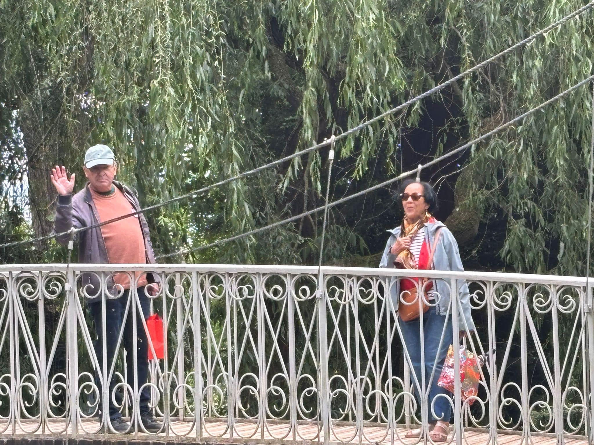 713724352_1291896093098214_4955939833643740718_n Two people stand on a decorative metal bridge railing; the man waves at the camera, the woman wears sunglasses nearby, with weeping willow branches behind them.