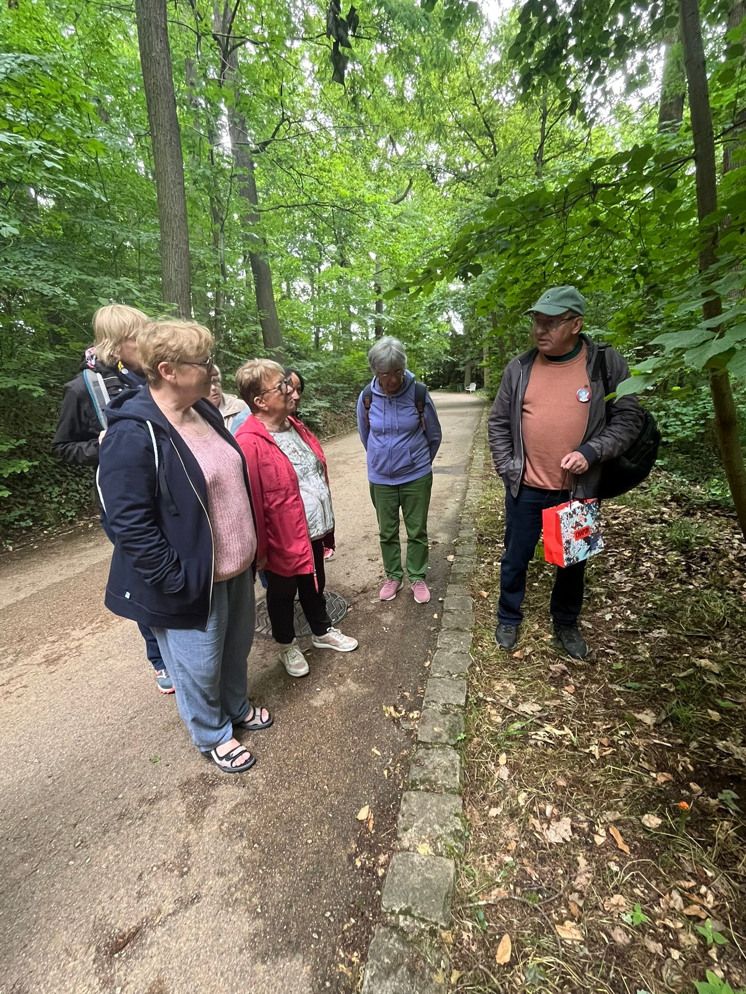 714343569_1291535003134323_8927031725104024479_n Group of adults standing on a tree-lined park path, chatting among themselves with backpacks visible