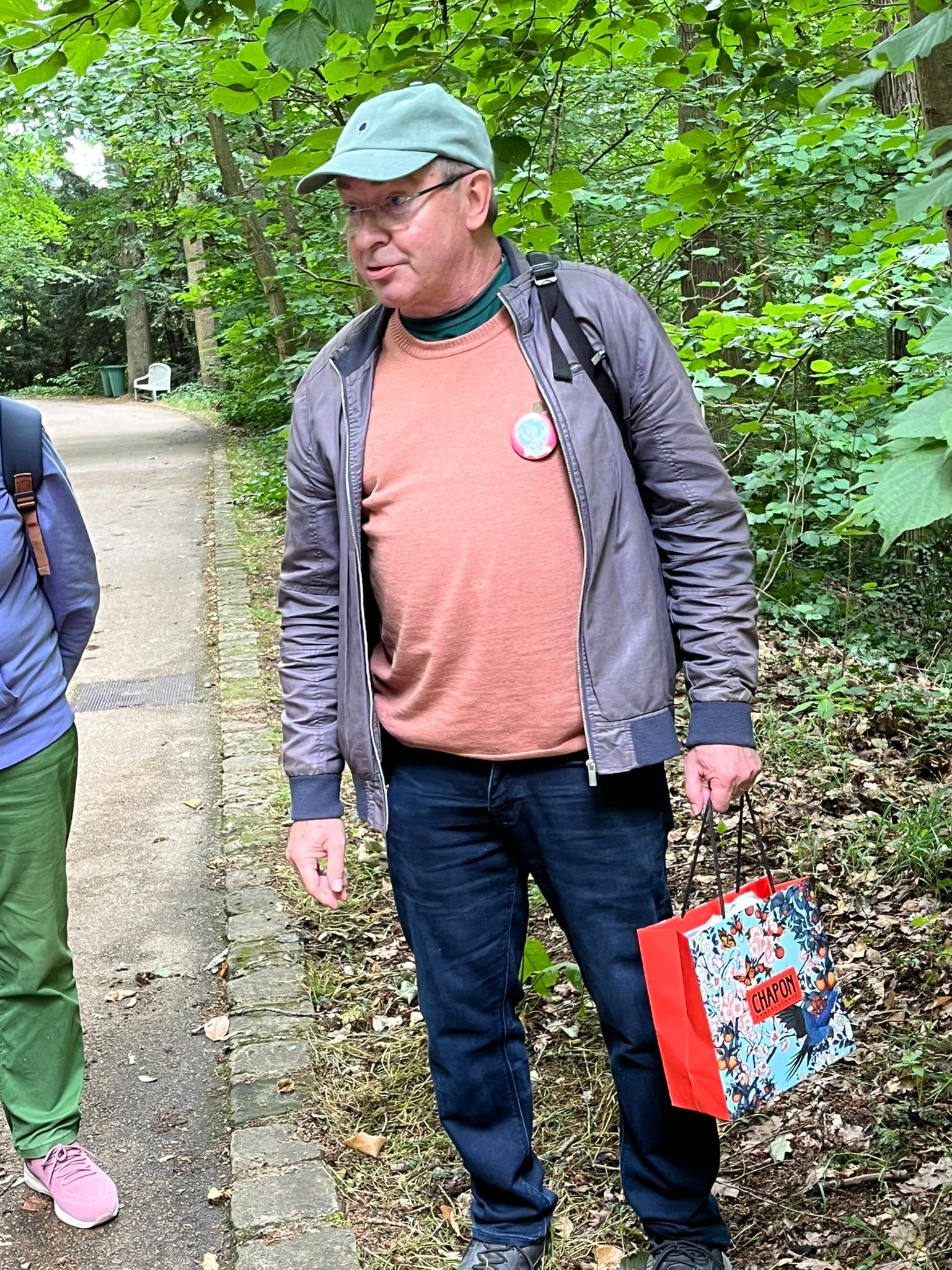 714759259_1291534649801025_9038023176005741443_n Man wearing a light blue cap, glasses, orange sweater and gray jacket stands on a park path with green foliage in the background, holding a colorful shopping bag.