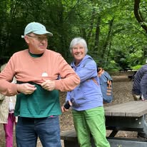 714839448_1291533503134473_7499757647287096951_n Man wearing a cap and peach shirt stands beside a smiling woman in a purple jacket near a wooden picnic table in a leafy park setting.