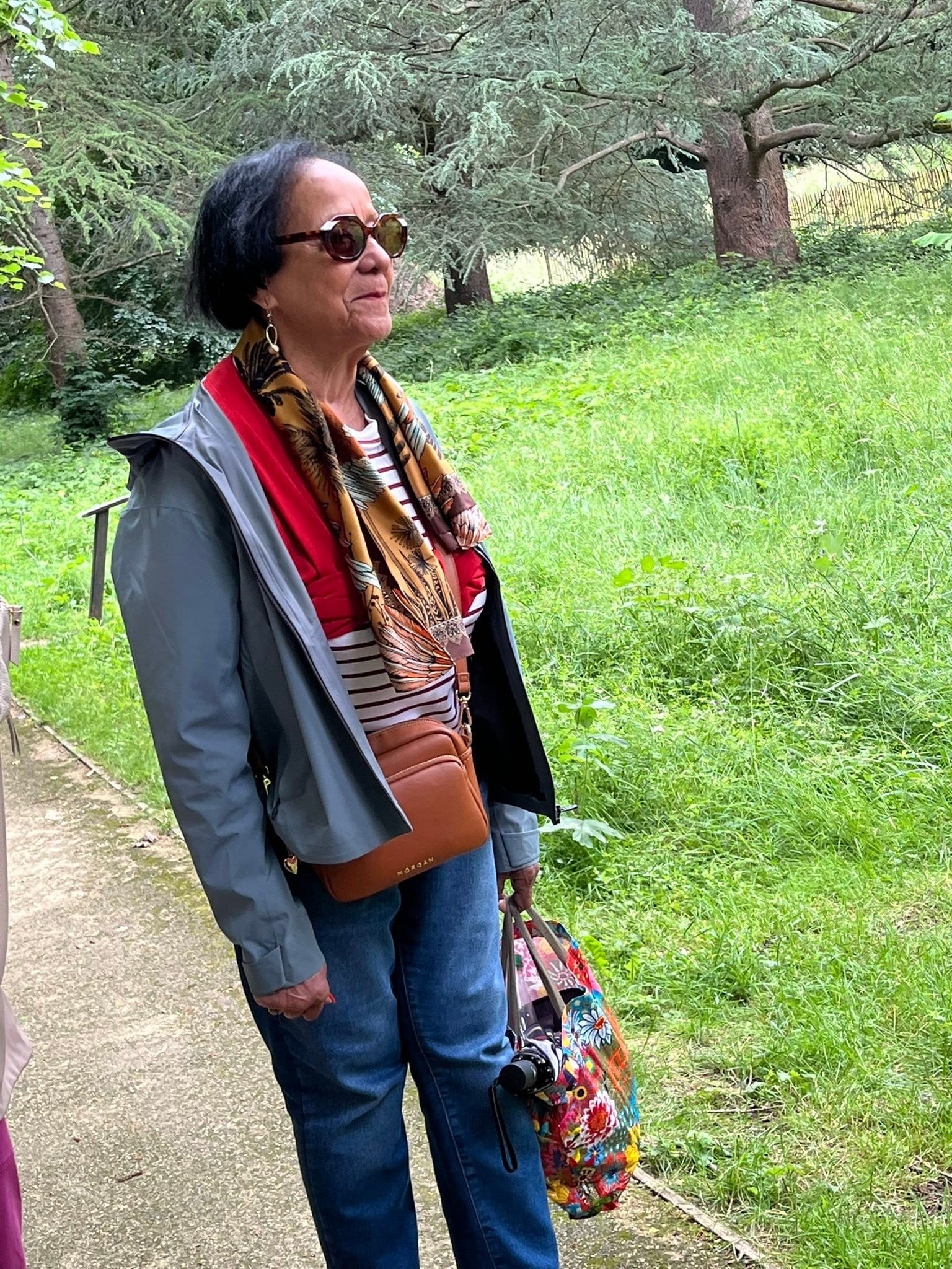 715464110_1291895956431561_5599111736552122746_n Older woman with short dark hair and large sunglasses stands on a park path, wearing a scarf and gray jacket.