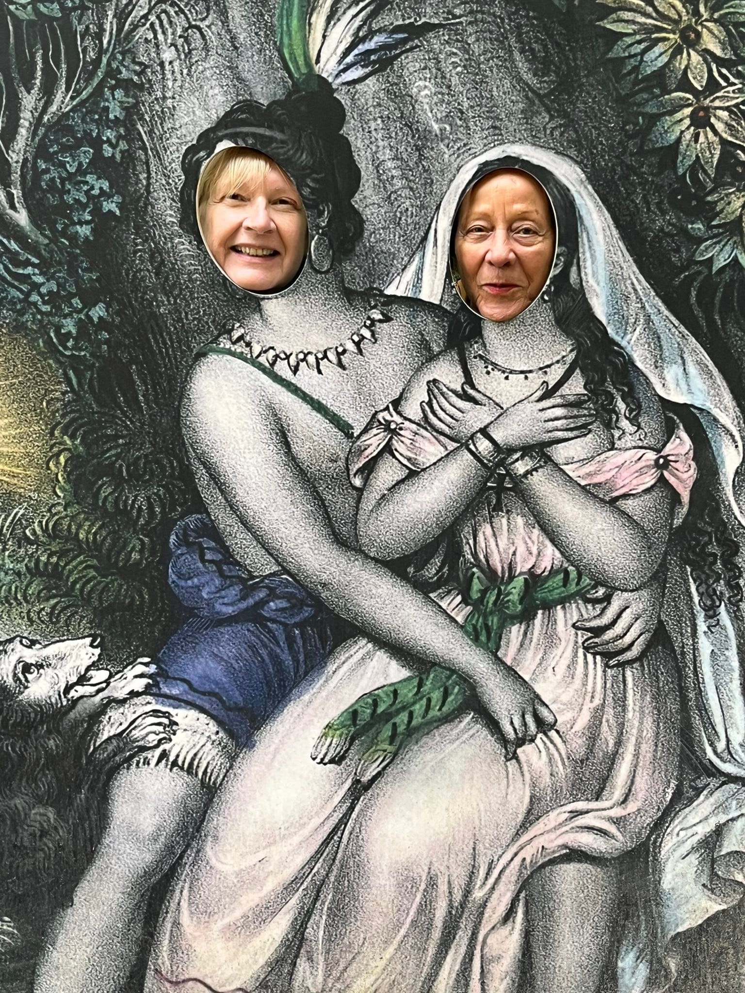 715790195_1291534173134406_3002370486484596565_n Two smiling women pose through face holes in a theatrical mural of two entwined figures in a forest scene, peeking from cutouts.