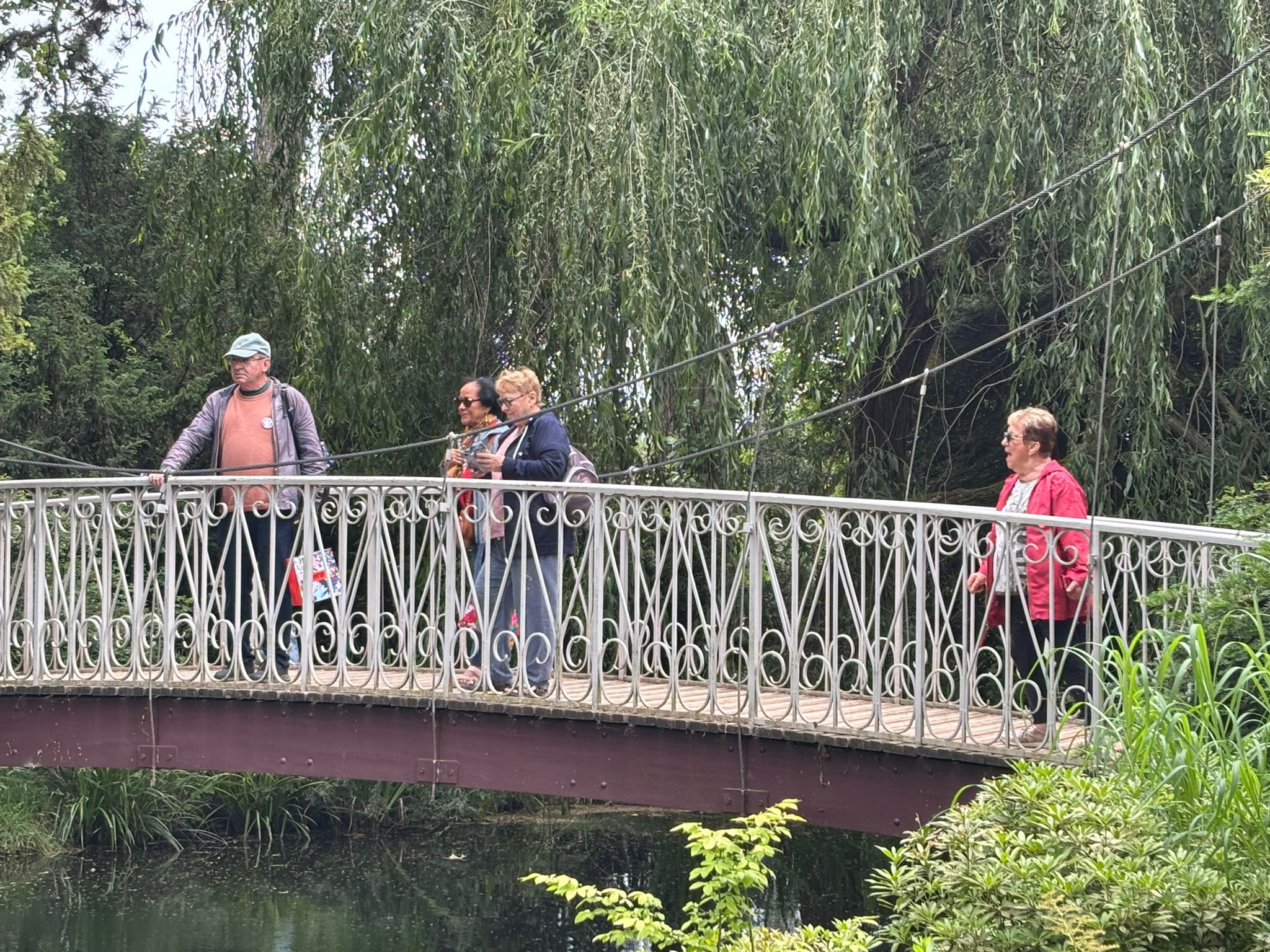 716570142_1291896219764868_3840522804349534064_n Several people cross a decorative iron bridge above a narrow waterway, with overhanging willow branches in the background.