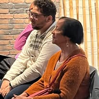 720177934_1297871859167304_4684972140375750656_n Three people sit on chairs indoors near a brick wall; a woman in an orange top sits in front, with a man in a striped shirt beside her, and a person in pink partly visible on the left.