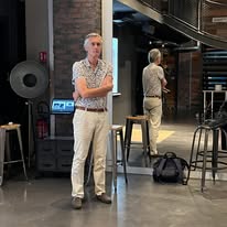 721429953_1297871815833975_6715638250128699945_n Man in a patterned shirt and light pants stands with arms crossed in a backstage rehearsal area beside a pillar.