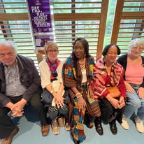 721575445_1298606972427126_7867633039707203782_n Six older adults seated on a wooden bench in a bright indoor space, smiling at the camera with a purple banner in the background indicating an event or program behind them.