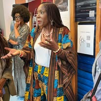 721667605_1298606632427160_2407583143983691725_n Woman in a colorful patterned coat speaks with animated hand gestures to a small group in a public space beside a blue vending machine/locker area.