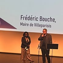721778427_1299411739013316_4105472646245846368_n Two speakers on a stage, a woman in a patterned dress and a man in a suit, with a backdrop reading 'Frédéric Bouche, Maire de Villeparisis'.
