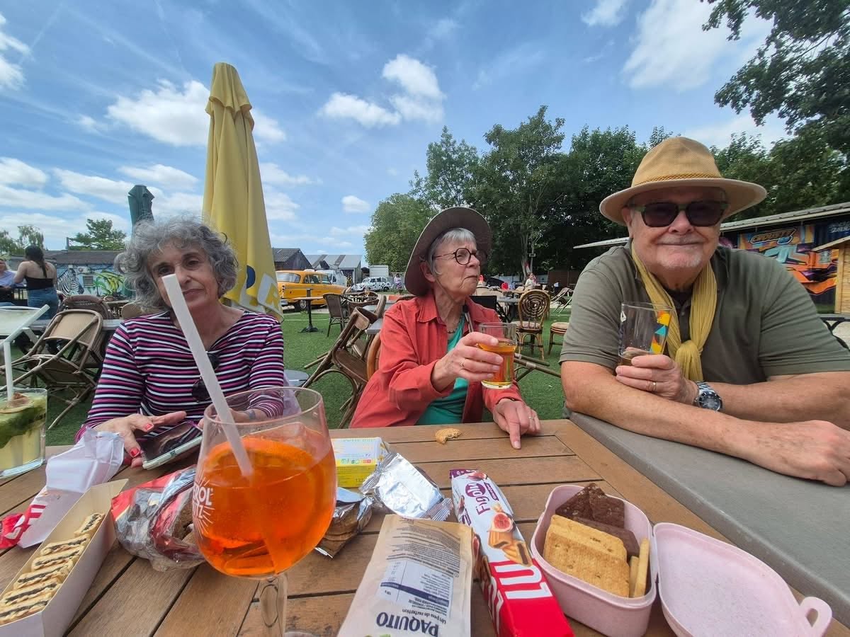 721995392_1302828072005016_2160757493672757776_n Three seniors seated at a wooden table outdoors, drinking and snacking on a sunny day at a casual cafe setting.