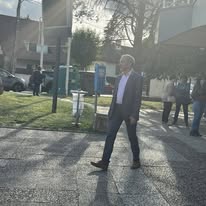 722336400_1299411772346646_4550887434567118554_n Man in a dark suit and tie walking on a sunlit sidewalk near office buildings.