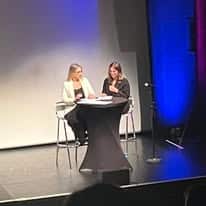 722447447_1299547992333024_1546169586362093797_n Two women sit at a tall round table on a stage, speaking into microphones during a panel discussion, with a white backdrop and blue stage lighting.
