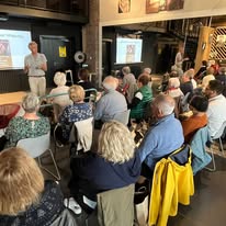 722909462_1297871799167310_450301903821461379_n Speaker presenting to a seated audience in a cafe-like room, with a projector screen behind and attendees listening attentively.