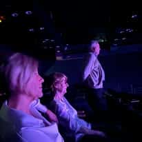 722953677_1299547835666373_2620055469649348095_n Audience seated in a dark theater with purple lighting; two women in the foreground and a man standing near the stage.