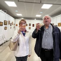 723428249_1299548138999676_6079583770721572250_n Two older adults in an art gallery pose for a selfie with a smartphone held up by the woman in a white jacket and beige bag.",