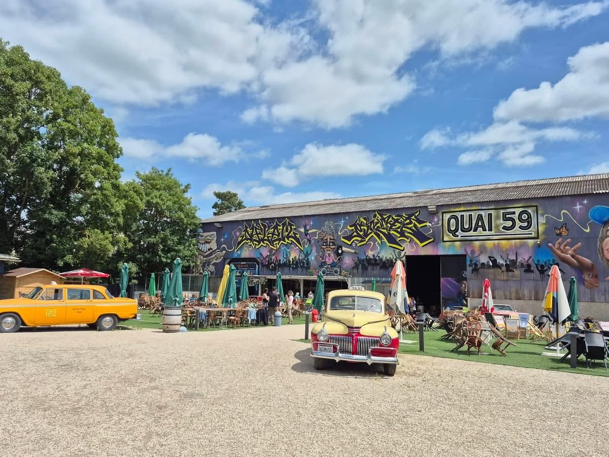 724484453_1302827945338362_7199909470620557317_n Vintage cars parked in front of a graffiti-covered building with an outdoor café and green umbrellas under a blue sky.