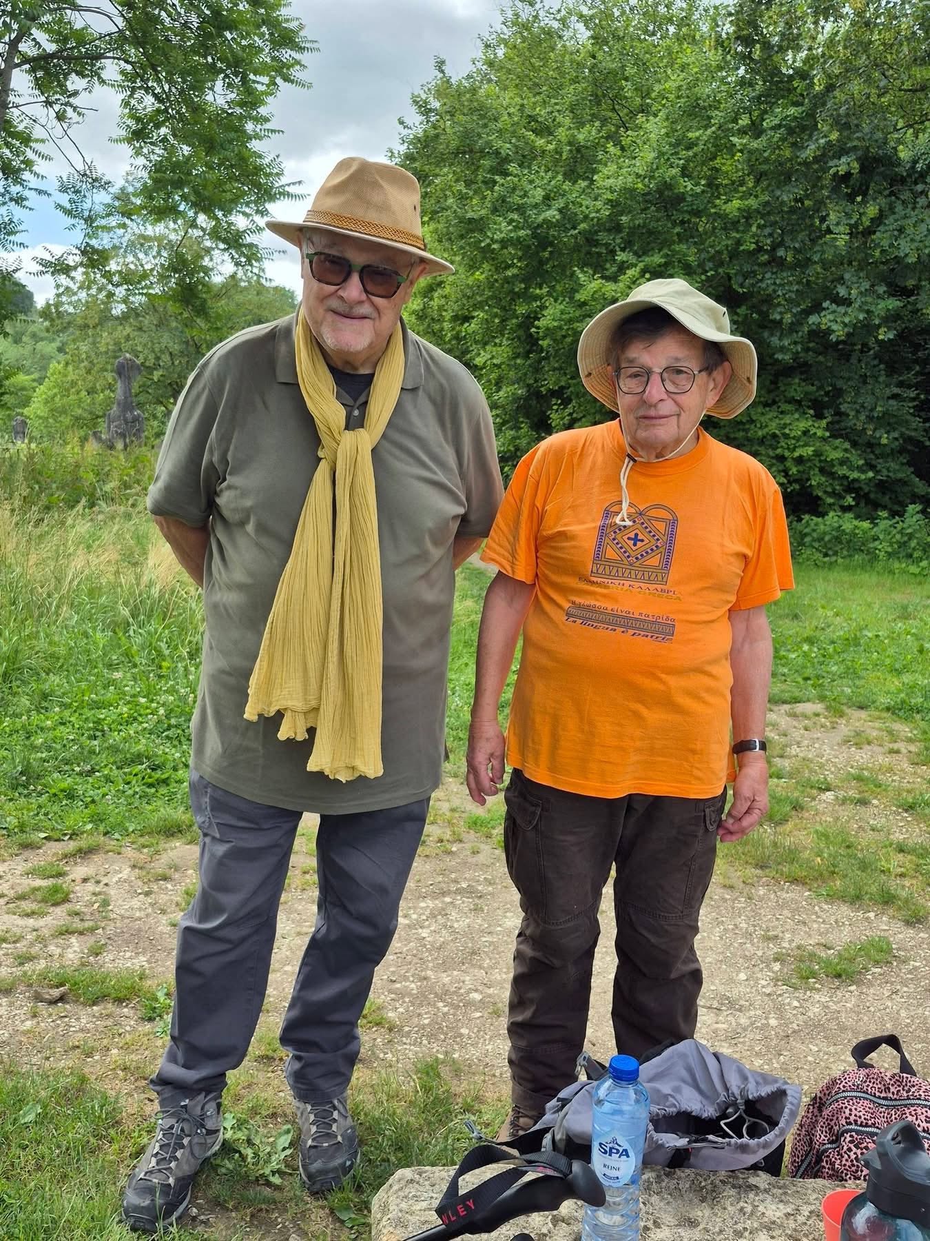 725633711_1302828038671686_2641368578752375557_n Two older men stand outdoors on a trail, wearing hats and hiking clothes, posing for a photo with greenery in the background.
