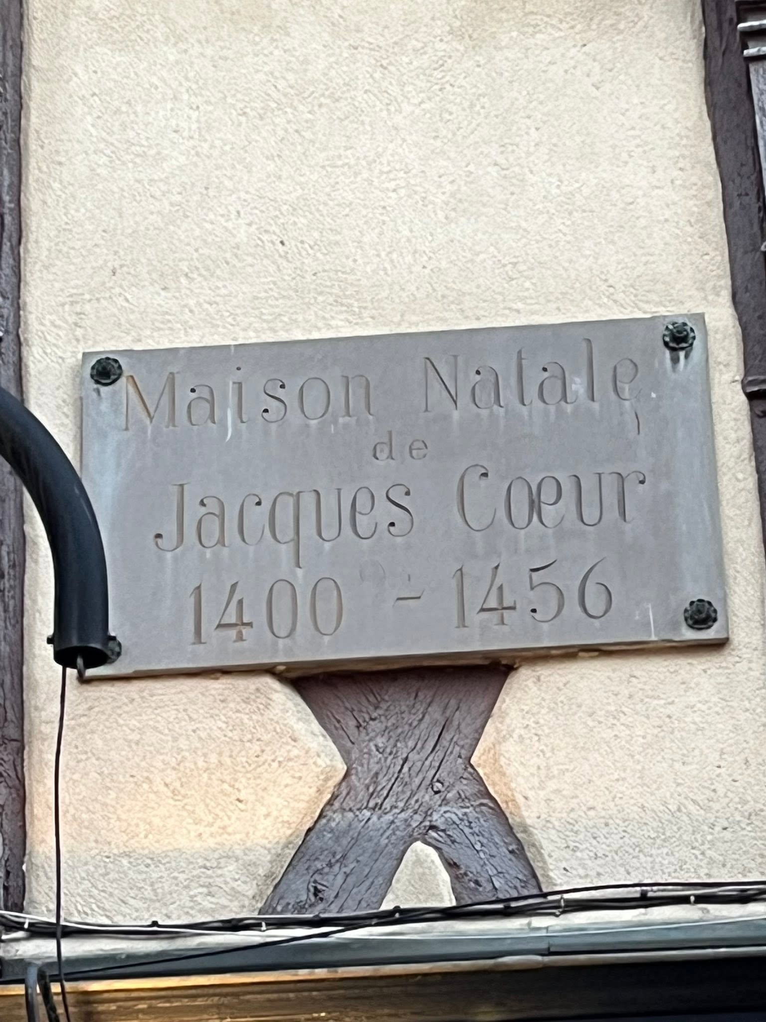 725879651_1305299908424499_393801974791887318_n Metal plaque mounted on a rough stone wall reading 'Maison Natale de Jacques Cœur', with the dates 1400–1456 and visible screws at the corners (adjacent cables near the plaque).