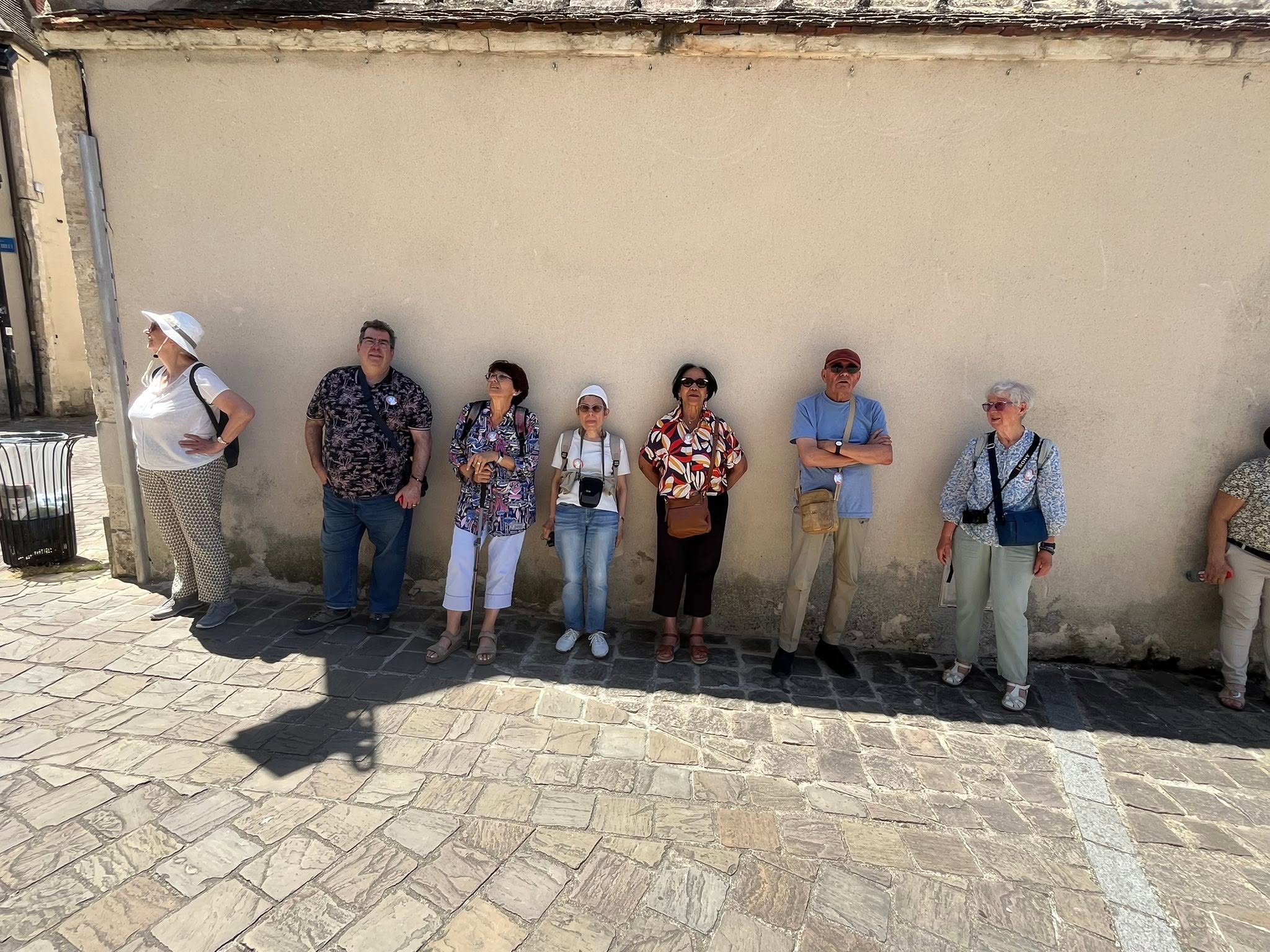 726427234_1305297978424692_3264973477585732135_n Eight seniors standing in a row along a sunlit beige wall on a cobblestone street, posing for a photo with casual clothing and bags/hat accessories.