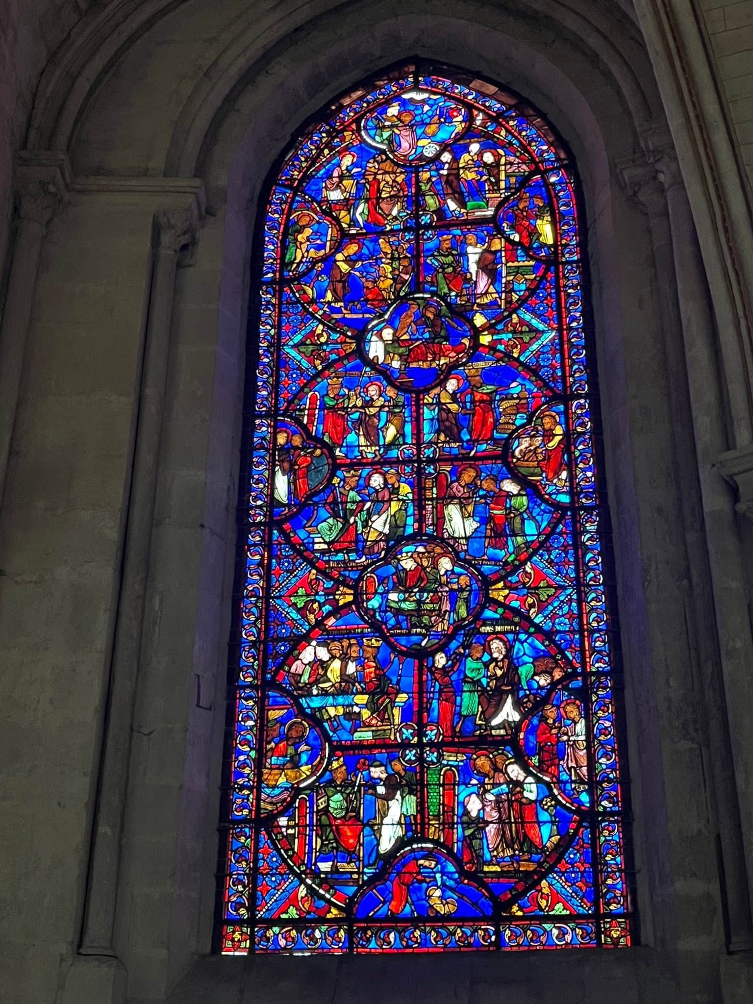 726470833_1305298898424600_5244402681992142980_n Colorful stained-glass window in a church, showing biblical scenes and figures in blue, red, and gold colors.