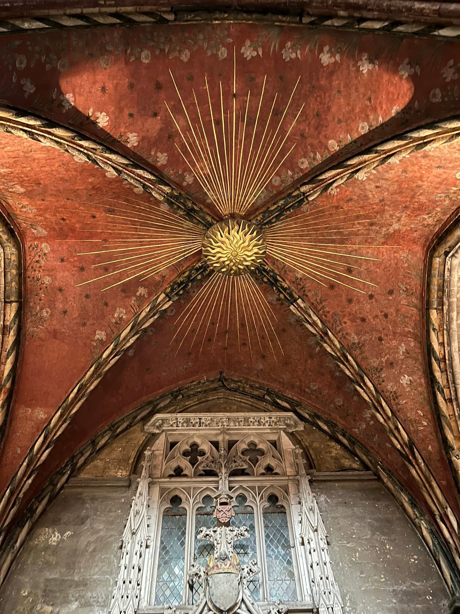 726575185_1305297911758032_2941830274773954131_n Gilded sunburst centerpiece on a red vaulted church ceiling with radiating gold rods and a carved stone window beneath.