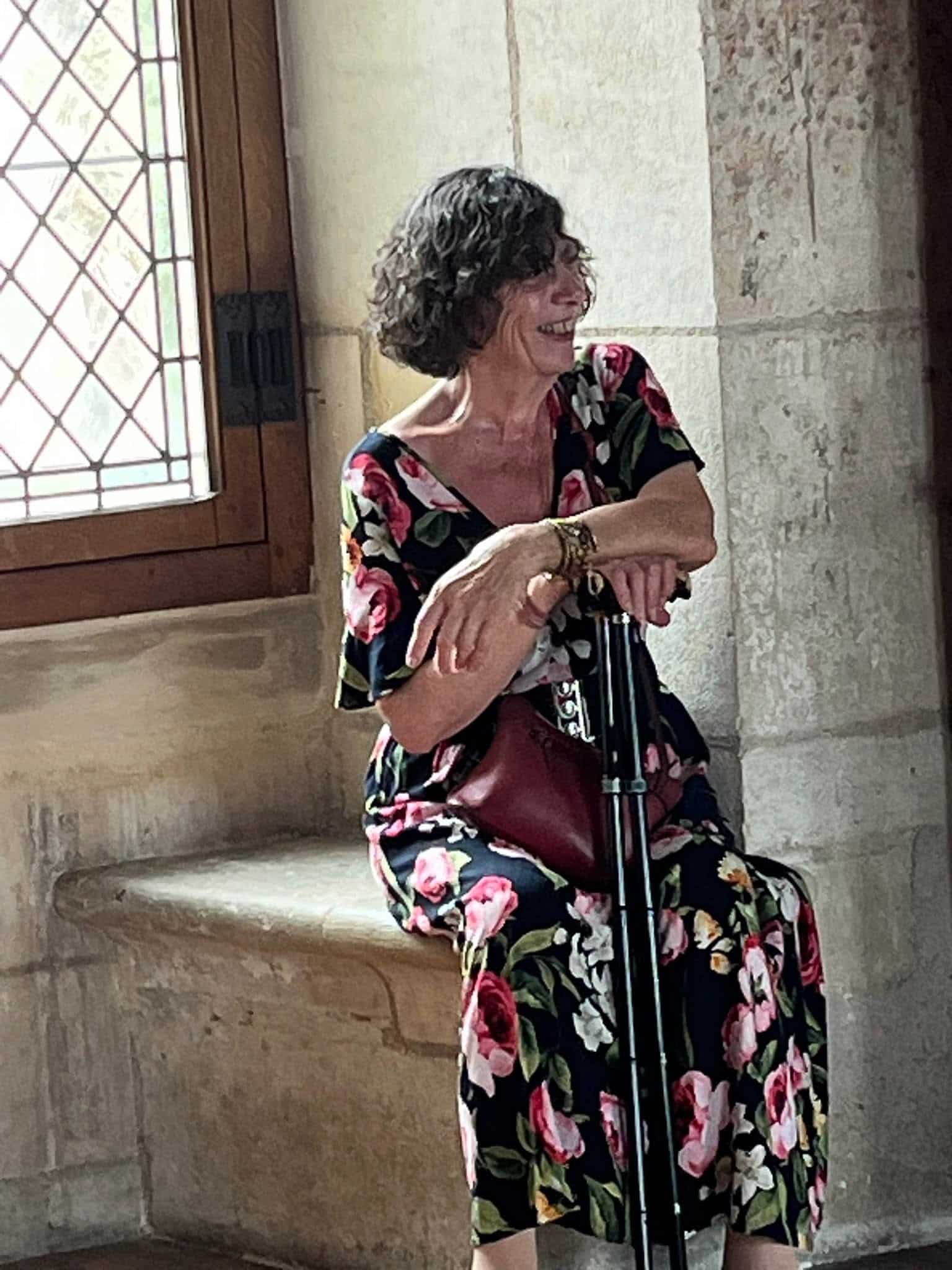 726576184_1306129631674860_2038508140267684373_n Smiling older woman with short curly gray hair wearing a pink floral dress sits on a stone ledge by a window in a historic building, resting her hands on a cane.