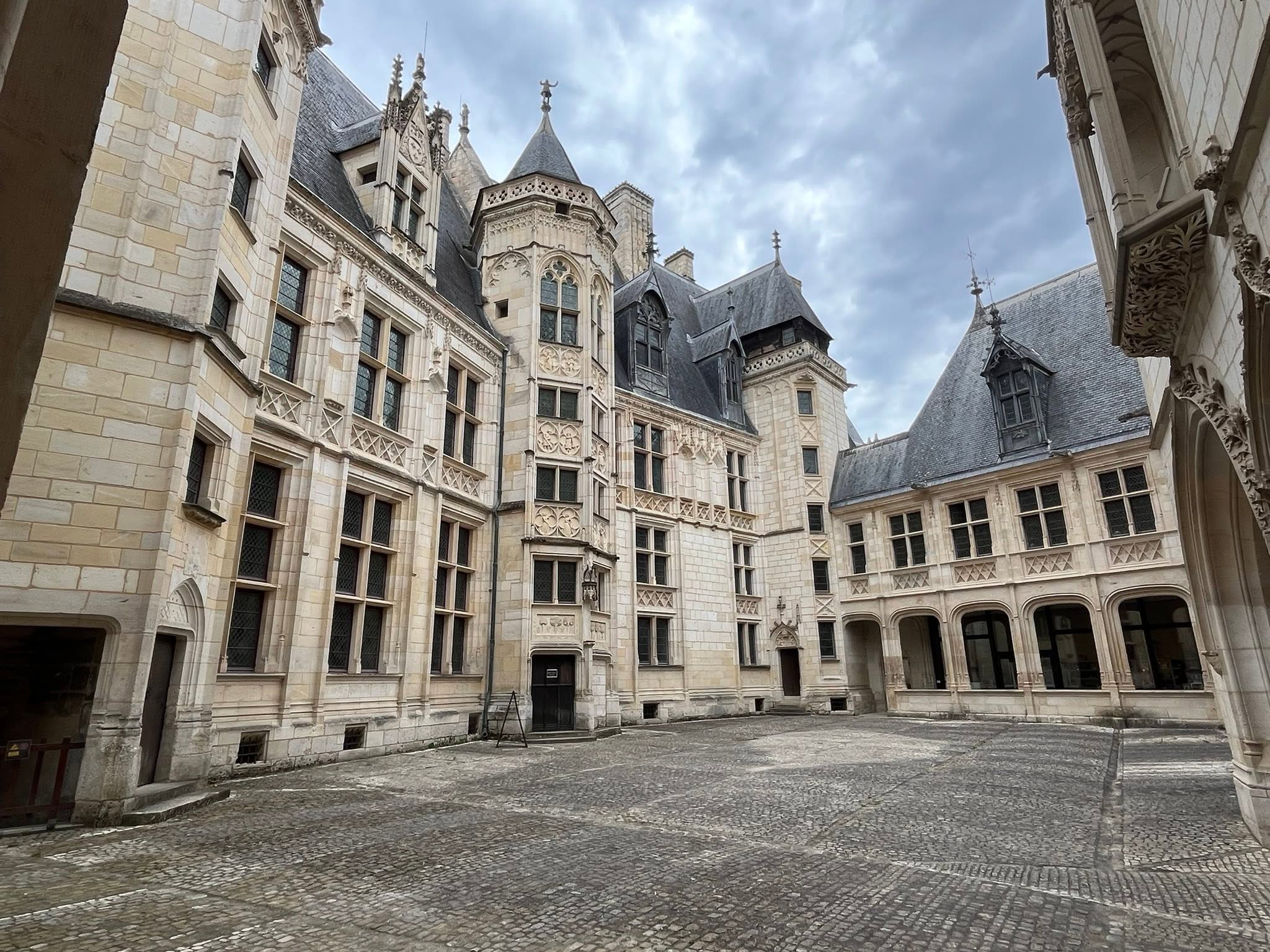 726576188_1306129595008197_2896267802388807786_n Historic stone courtyard with Gothic-inspired façades, pointed windows, and decorative carvings under a cloudy sky.