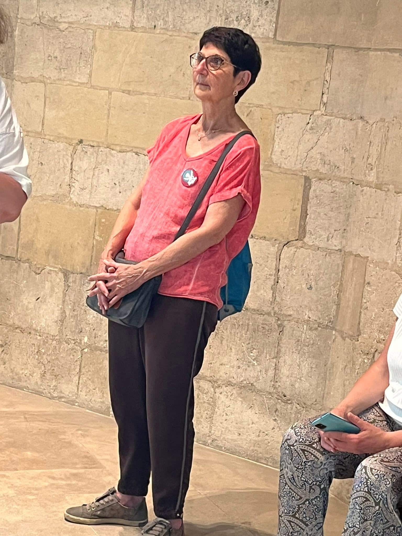 726576202_1306129278341562_3583077008267902993_n Older woman with short dark hair and glasses wearing a red blouse and black pants, standing against a stone wall with a crossbody bag.",