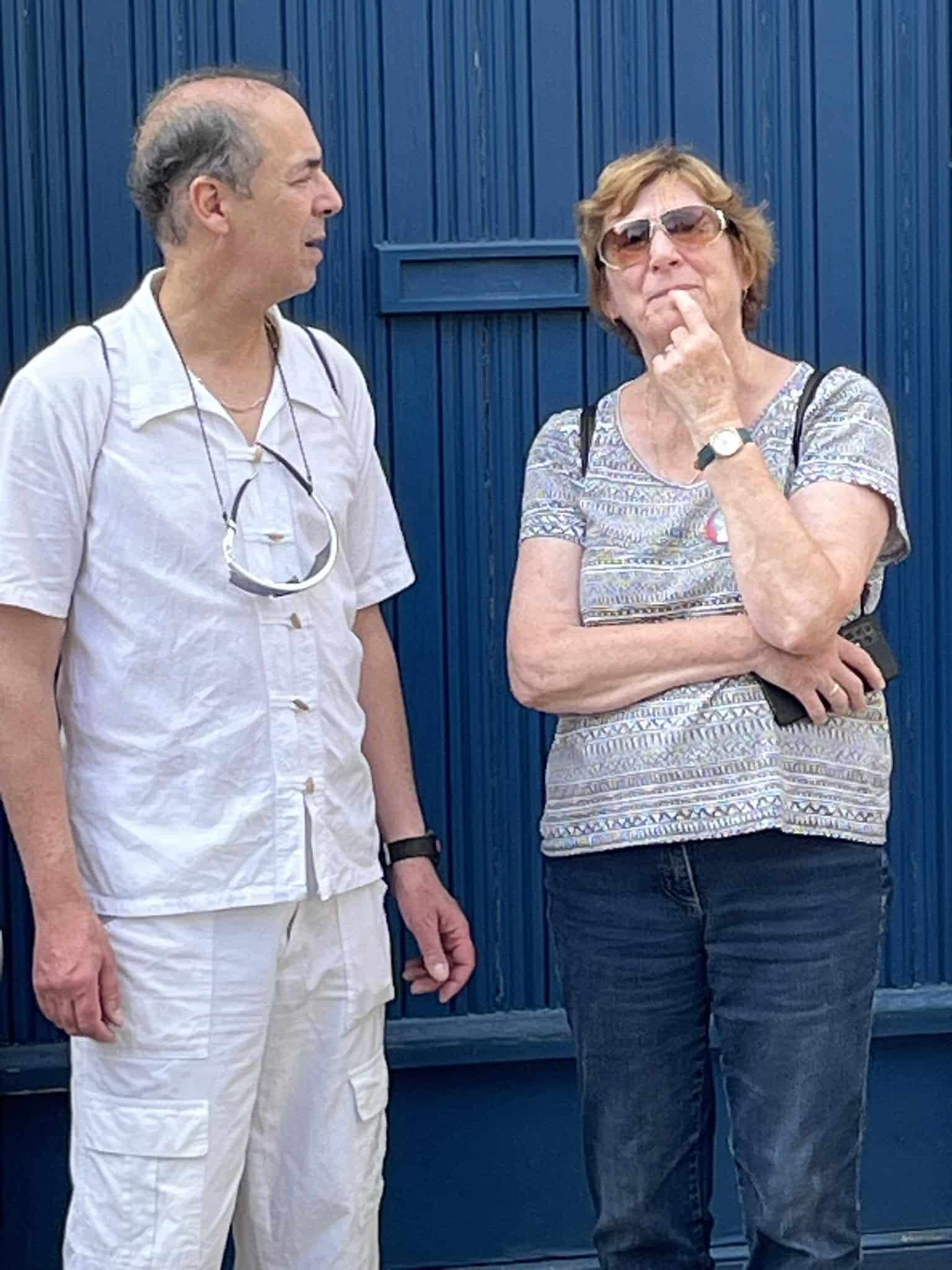 726607058_1305290978425392_2895419004001118278_n Two adults stand beside each other in front of a blue corrugated metal wall; man in a light shirt with sunglasses hanging from his chest, woman in a patterned top wearing sunglasses.