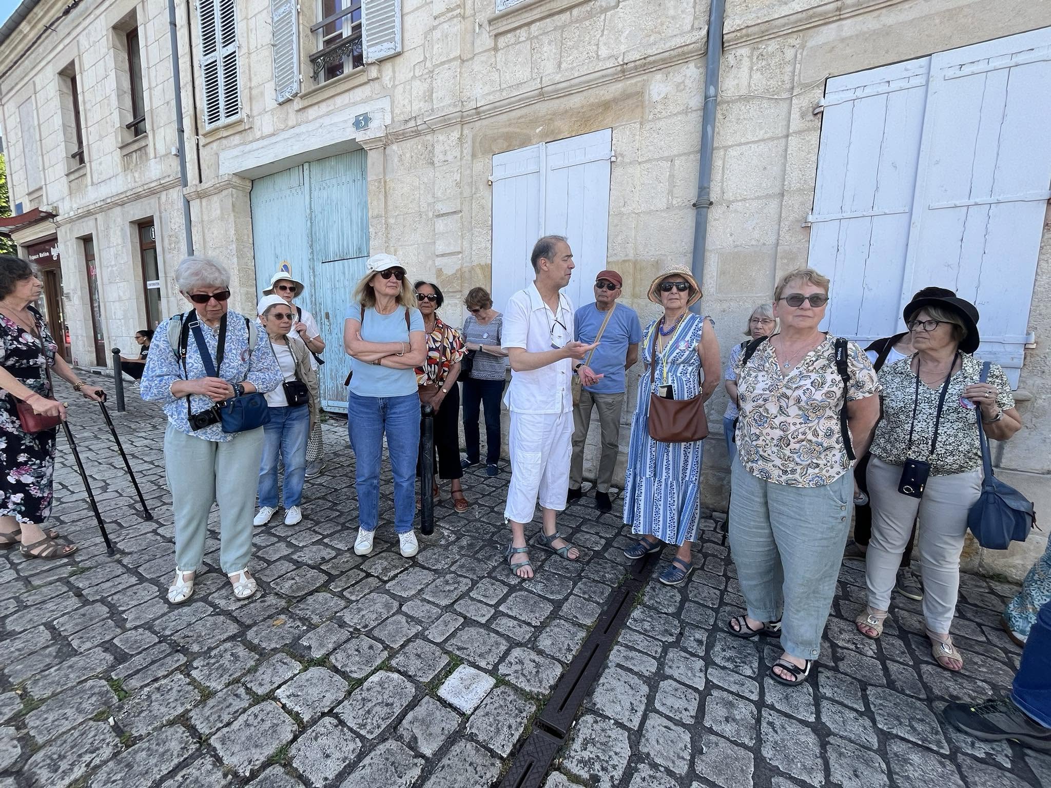 726607066_1305290948425395_1137921869085020363_n Group of older adults standing on a cobblestone street beside a stone building with blue shutters; a guide in white clothes gestures while talking to them.