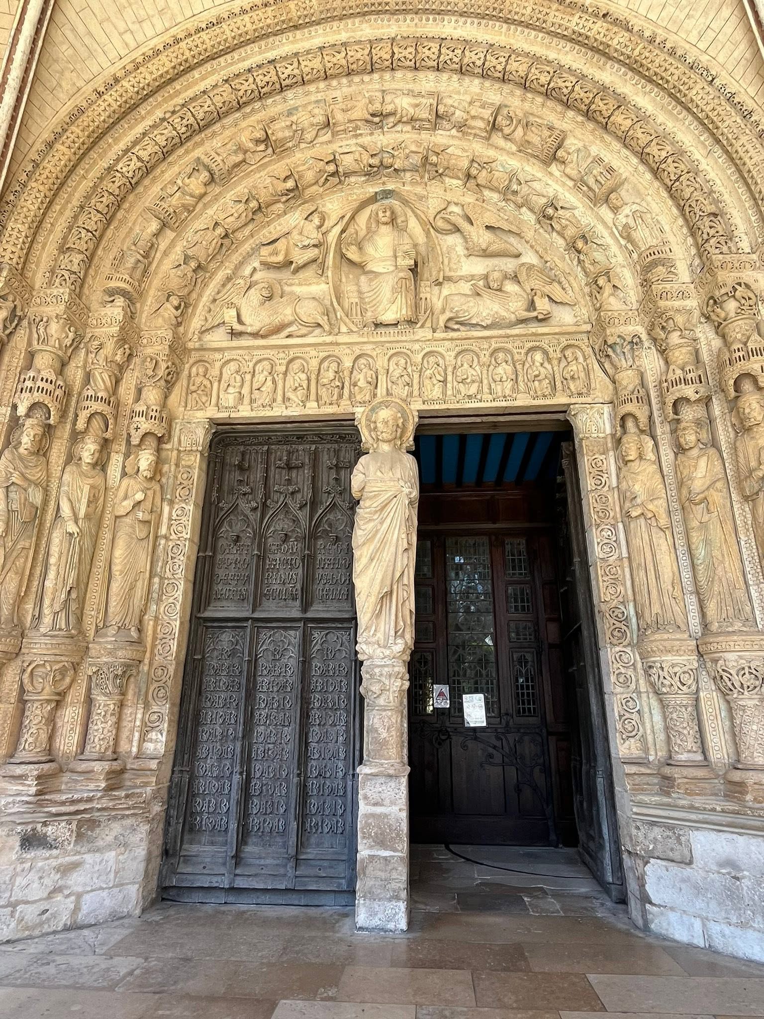 726636972_1305297715091385_1842046326293945308_n Ornate stone church portal with multiple detailed reliefs of saints and biblical scenes surrounding a central seated figure above heavy wooden doors.