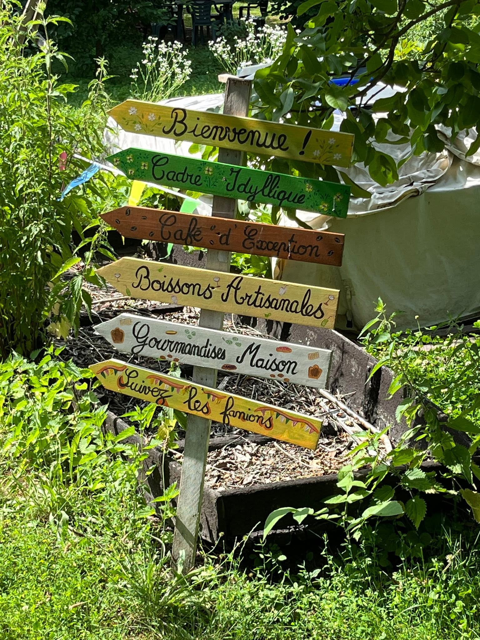 726717920_1305827525038404_7575506798371677789_n Colorful wooden signpost in a garden with French phrases like Bienvenue and Gourmandises Maison, inviting visitors.