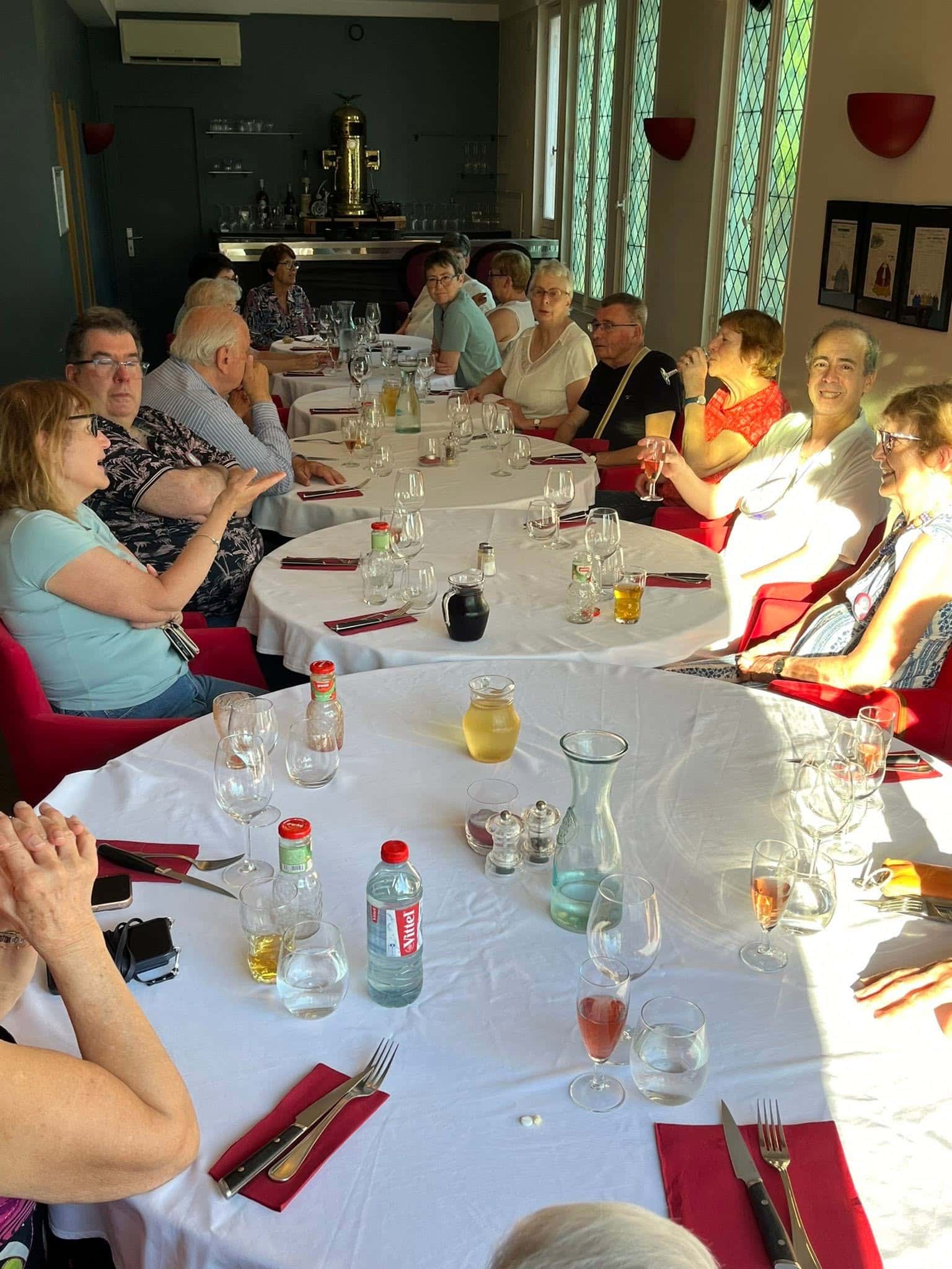726717940_1305299445091212_964347891086906871_n Group of seniors seated along a long, white-tablecloth dinner table in a sunlit dining room, chatting and enjoying drinks and food.