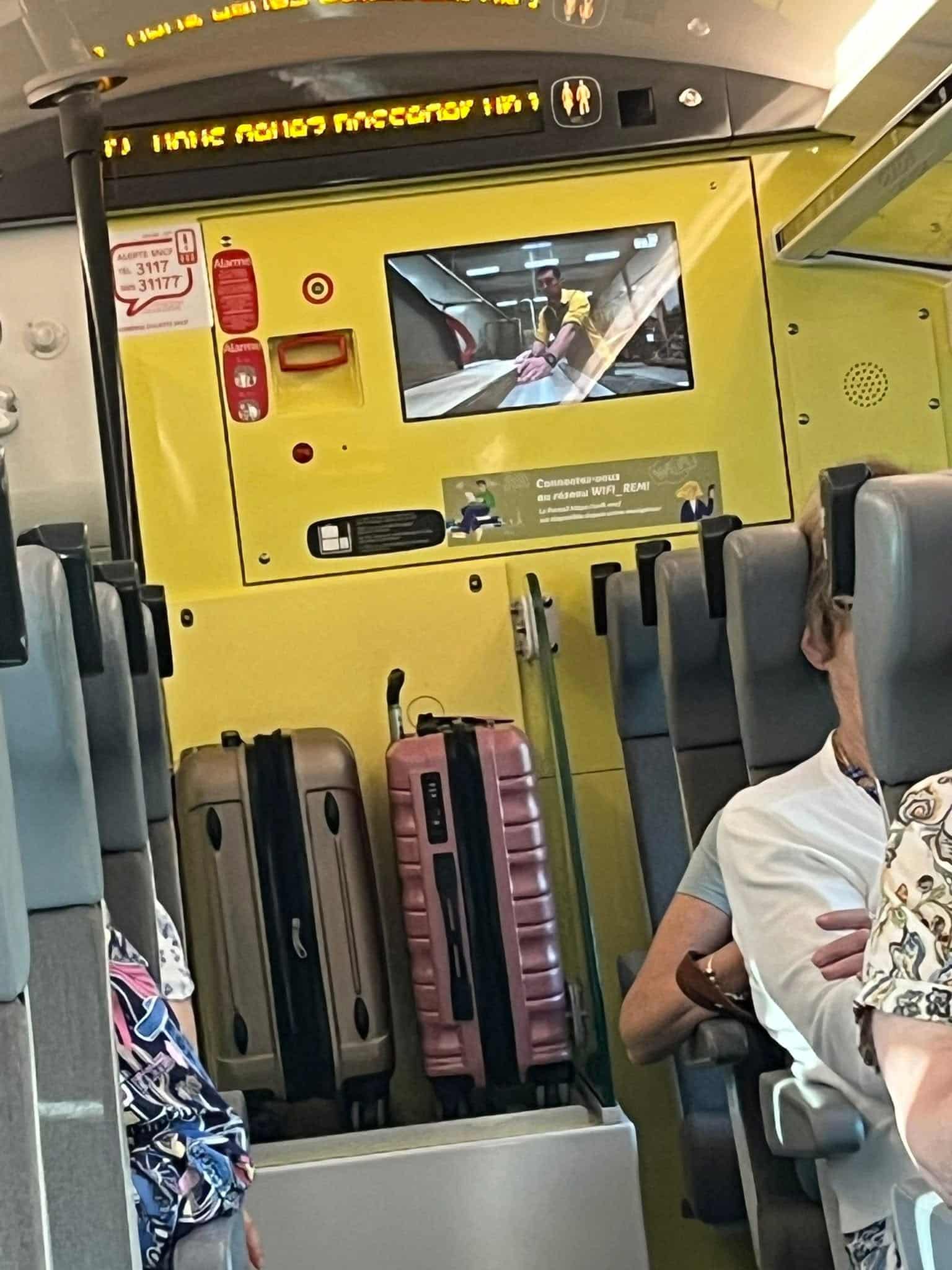 726733639_1304748618479628_3888442053680147007_n Interior of a yellow airplane cabin with rows of seats and luggage standing in the aisle; a screen shows a man in a yellow shirt.