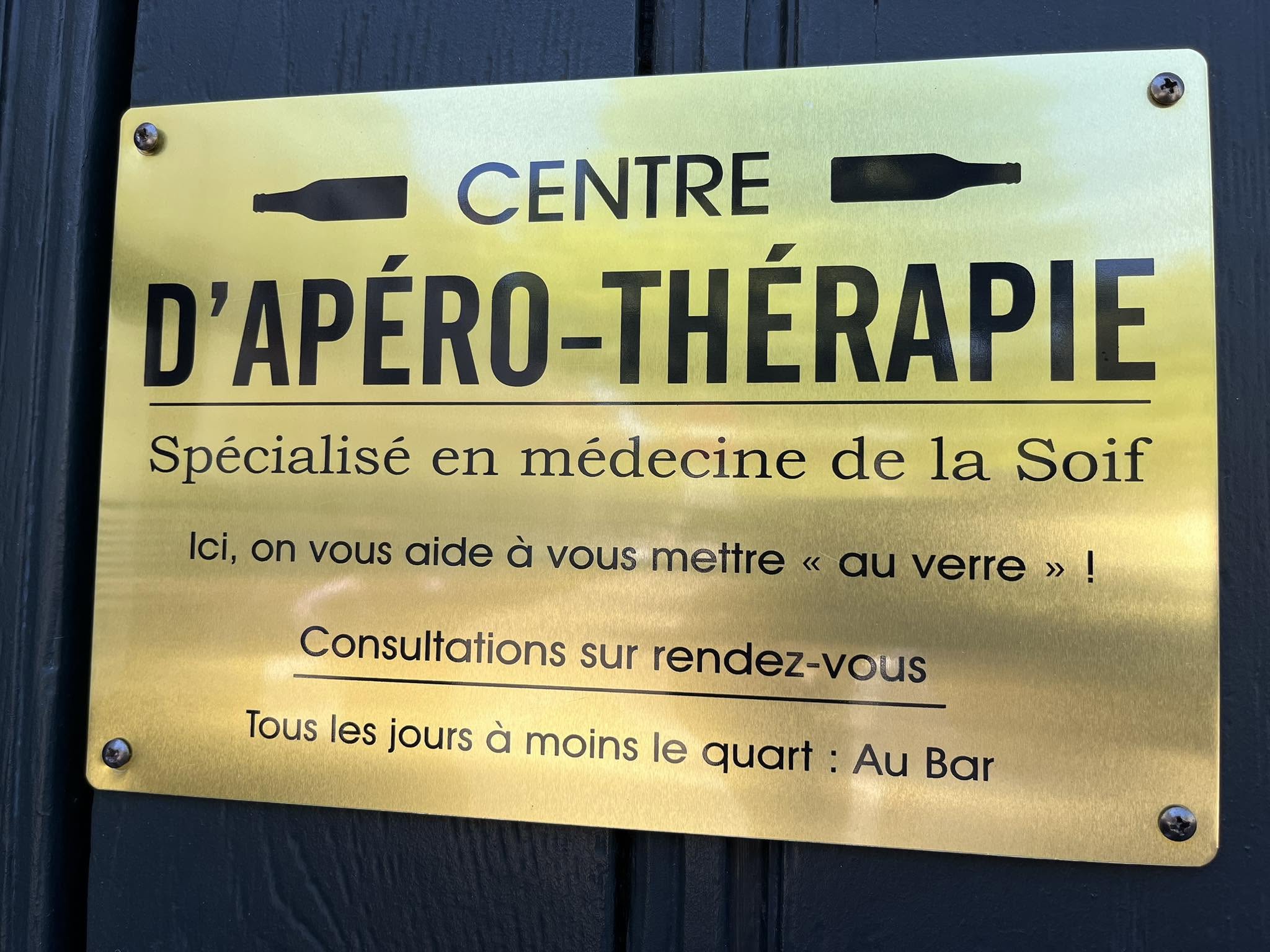 726900712_1305292181758605_1629272236981622820_n Gold sign reading 'Centre d'Apéro-Thérapie' with bottle silhouettes on each side, mounted on dark wood.