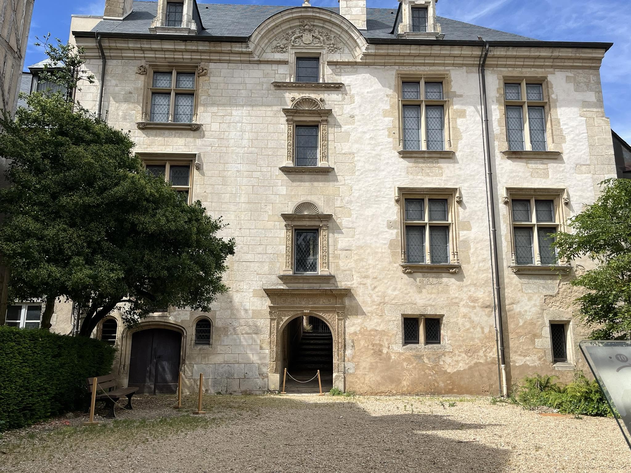 726914716_1305292565091900_826165010568874032_n Front view of a beige stone building with ornate window frames and a large arched doorway, flanked by green trees under a blue sky.