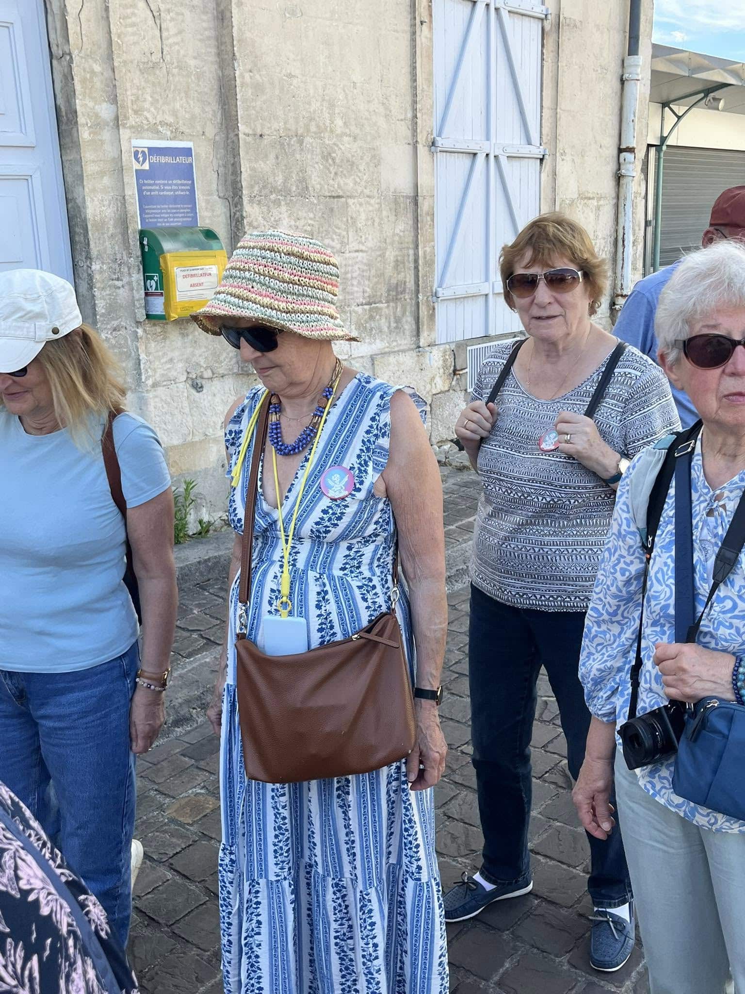 727059615_1305290258425464_7924145125192597739_n Group of older women walking along a stone street, wearing sun hats, sunglasses, and summer outfits with badges.