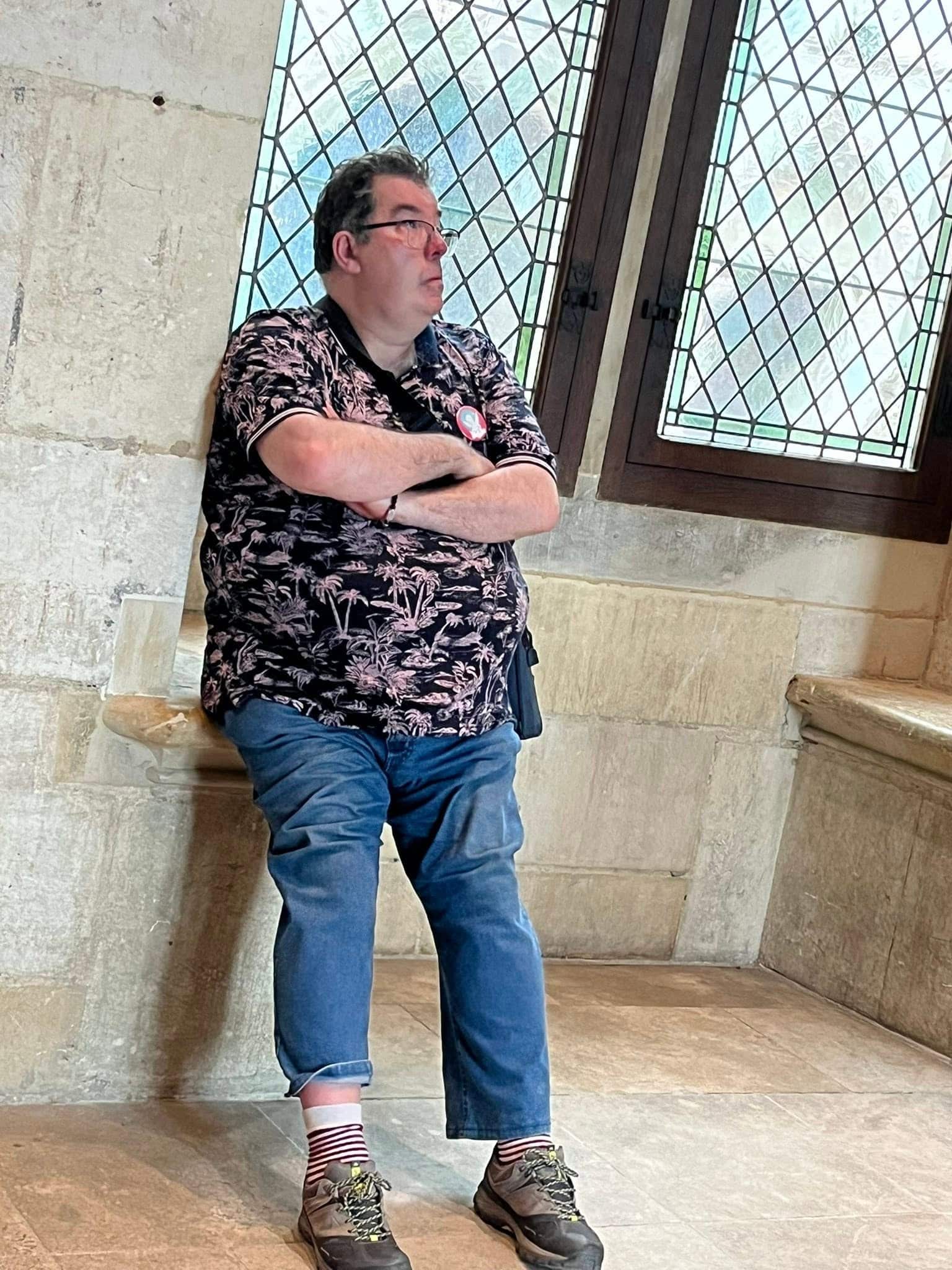 727059805_1306129485008208_1189994420918962226_n Man with arms crossed wearing a floral shirt, jeans, and striped socks stands in a stone-walled room with a diamond-pattern window behind him.