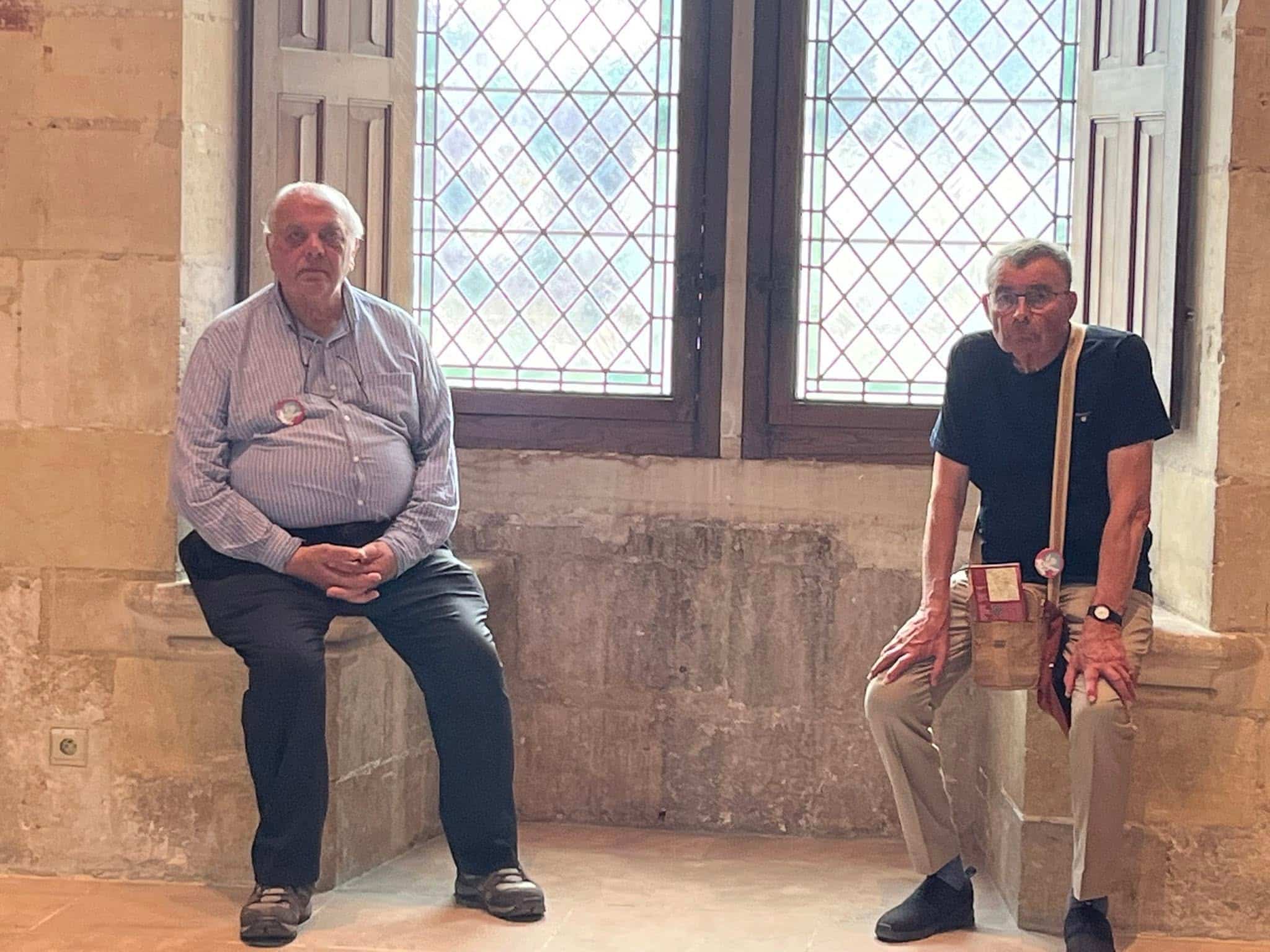 727158486_1306130145008142_2628975179849872017_n Two men sitting on a stone windowsill in a historic room, with a diamond-pane window behind them; both wear badges.